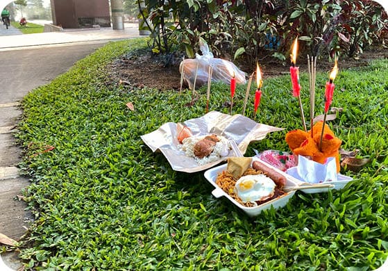 Singapore Sep2021 Packed food, burning candles and joss sticks incense placed on grass in neighbourhood heartland as offerings during 7th month Hungry Ghost Festival. Concrete burner in background.